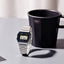 Digital watch next to a black mug on a light surface